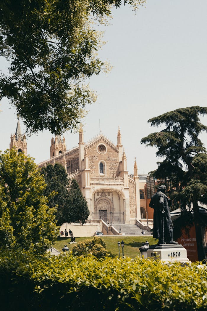 View of the San Jeronimo el Real Church, a historic landmark in Madrid, Spain, surrounded by lush trees and statues.